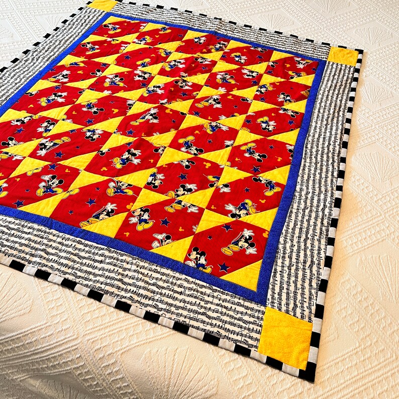 May include: A handmade quilt featuring a repeating pattern of Mickey Mouse on a red background, interspersed with yellow diamond shapes. The quilt has a blue border and a black and white striped edge.