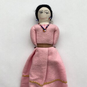May include: A handmade fabric doll with black hair, a pink dress, and a beaded necklace. The doll has painted eyes and wears earrings.