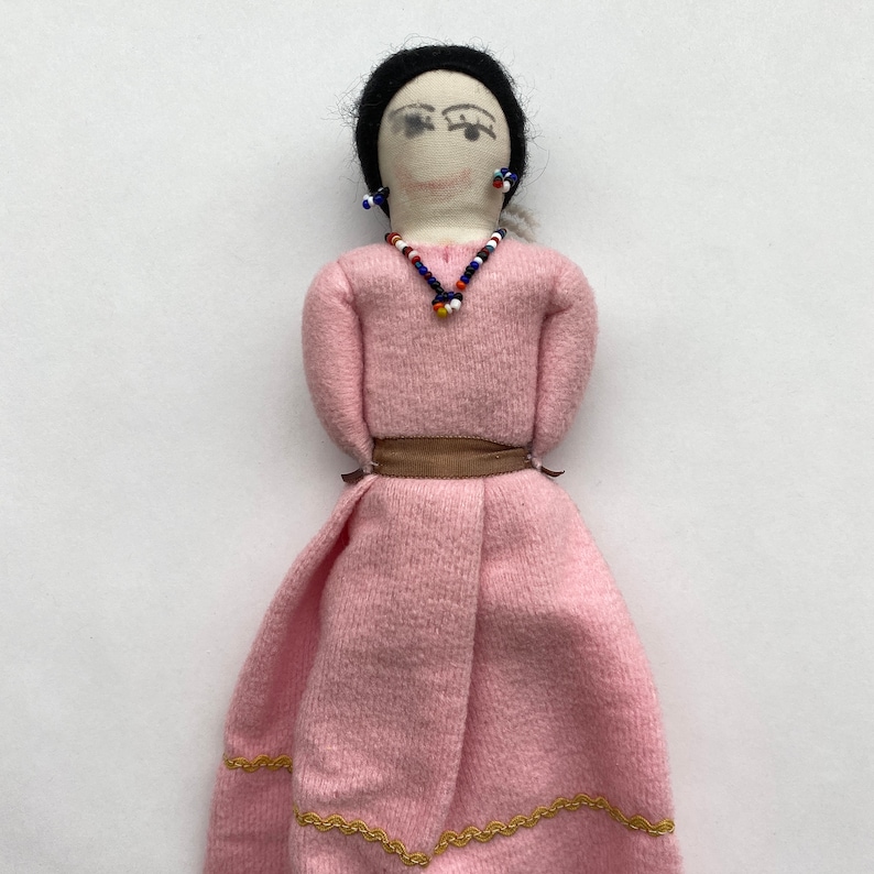May include: A handmade fabric doll with black hair, a pink dress, and a beaded necklace. The doll has painted eyes and wears earrings.