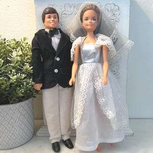 May include: A vintage wedding doll set featuring a groom in a black tuxedo with a white shirt and bow tie, and a bride in a white lace wedding dress with a veil. The dolls are standing side by side, showcasing a classic bridal theme.