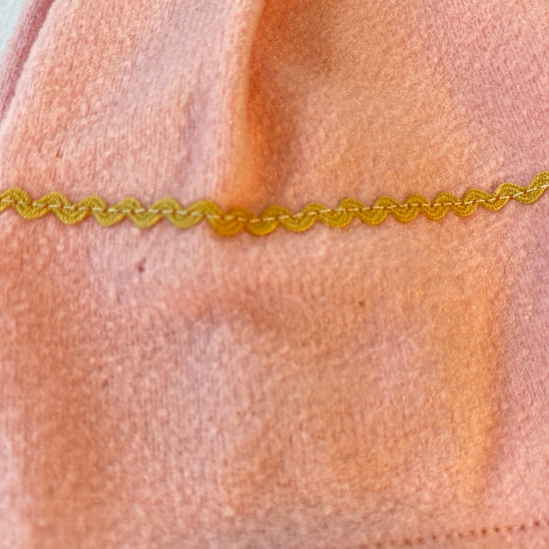 May include: A close-up of a pink fabric with a gold scalloped trim. The fabric has a soft, fuzzy texture.
