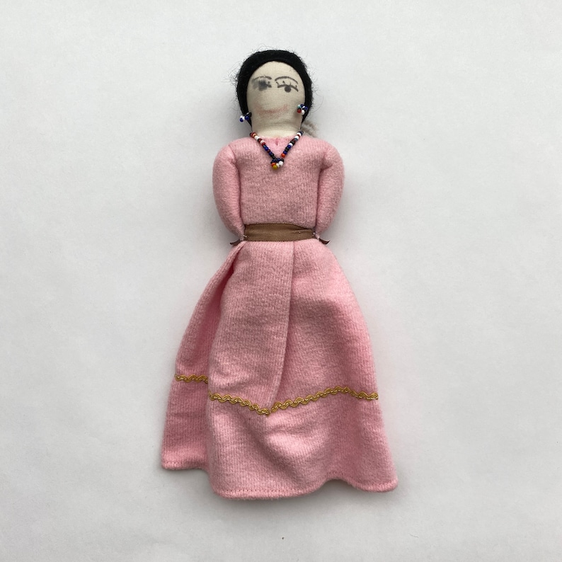 May include: A handmade fabric doll wearing a pink dress with gold trim and a brown belt. The doll has black hair, a painted face, and beaded earrings and necklace.