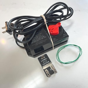 May include: A black Lionel transformer with a red power dial, a black power cord, and a separate black Lionel CTC Lockon track connector with metal clips. A thin green wire is coiled next to the connector, and the transformer is tied with a rubber band.