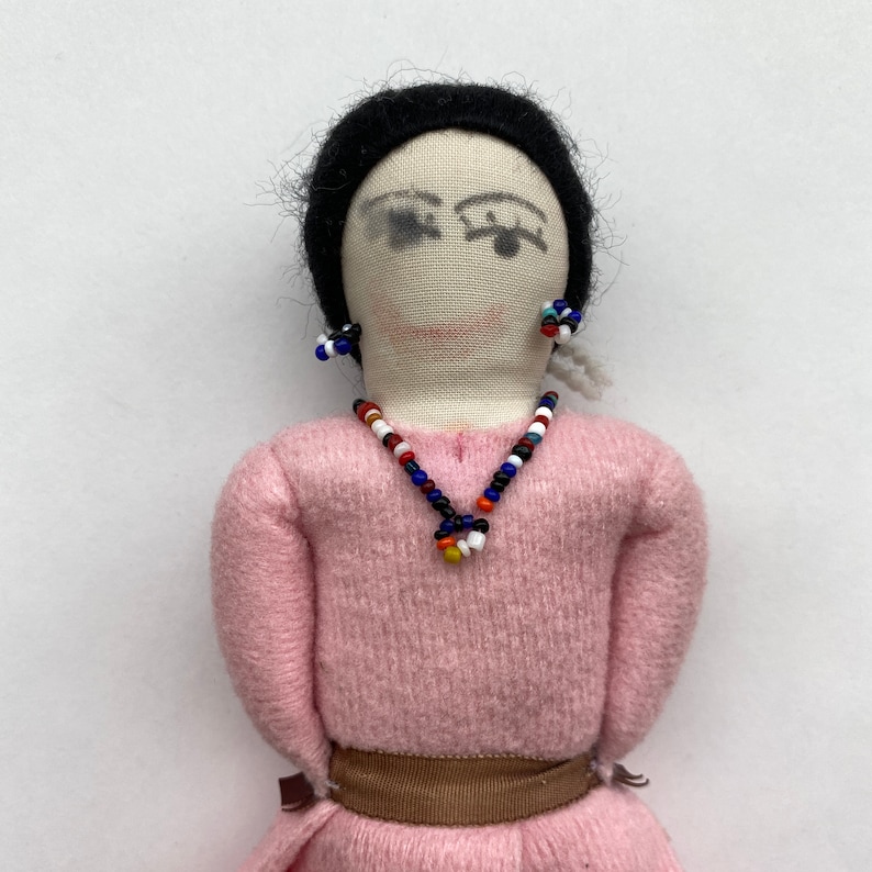 May include: A handmade doll with black hair, a pink sweater, a brown belt, and a beaded necklace and earrings.