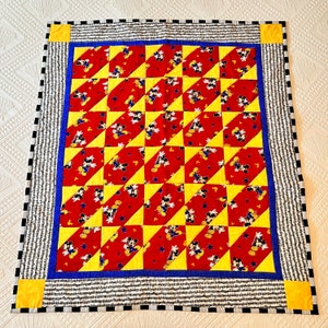 May include: A handmade quilt featuring a repeating pattern of Mickey Mouse on red fabric, alternating with yellow triangles. The quilt has a blue border, a black and white striped border, and yellow corner squares. The quilt is square-shaped.