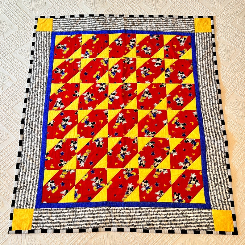 May include: A handmade quilt featuring a repeating pattern of Mickey Mouse on red fabric, alternating with yellow triangles. The quilt has a blue border, a black and white striped border, and yellow corner squares. The quilt is square-shaped.