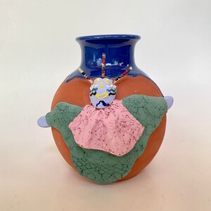 May include: A terracotta vase with a blue top and a whimsical clay figure attached. The figure has a blue face with yellow and black details, a pink and green patterned cape, and blue arms.