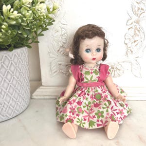 May include: A vintage doll with brown hair and blue eyes wearing a pink and green floral dress. The doll has a pink ribbon in her hair and a silver necklace. The doll is sitting on a white surface.