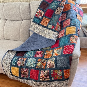 May include: A patchwork quilt with a variety of colorful fabrics in shades of red, orange, blue, green, and brown. The quilt is made up of squares and rectangles, and it has a paisley border. The quilt is draped over a gray couch.