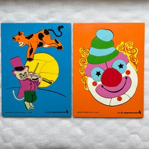May include: Two vintage wooden jigsaw puzzles. One features a cat playing a violin with a cow jumping over a yellow ball, on a blue background. The other puzzle is a clown face with a red nose and blue and green hair, on an orange background.