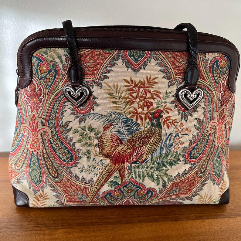 以下が含まれることがあります： ペイズリーとキジのプリントが施された、ブラウンレザーとキャンバスのトートバッグ。バッグには、ハート型のメタルアクセントが付いた、編み込みレザーのハンドルが2つ付いています。