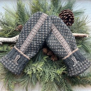 May include: A pair of gray and brown knitted mittens with a diamond pattern and a decorative stripe down the center. The cuffs are dark gray with a silver boot charm. The mittens are displayed on a bed of pine needles and pine cones.