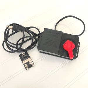 May include: A black Lionel train transformer with a red control handle, a black power cord with a two-prong plug, and a separate black Lionel CTC Lockon track connector. The transformer casing features molded text including caution warnings and model details.