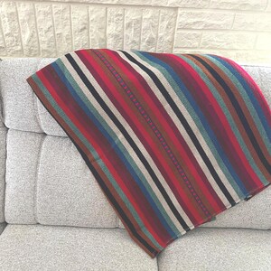 May include: A multi-coloured striped blanket, draped over a grey sofa. The blanket has vertical stripes in red, blue, green, black, and tan. The sofa is upholstered in a textured, light grey fabric.