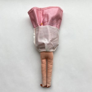 May include: A handmade doll with pink and white fabric clothing. The doll has long legs and is standing on a white surface.