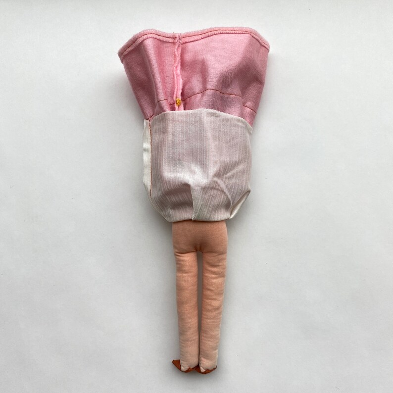 May include: A handmade doll with pink and white fabric clothing. The doll has long legs and is standing on a white surface.