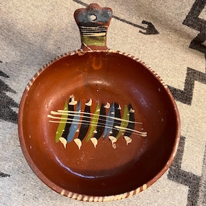 May include: A terracotta bowl with a handle, painted with a geometric design in black, blue, green, and white. The bowl is approximately 8 inches in diameter.