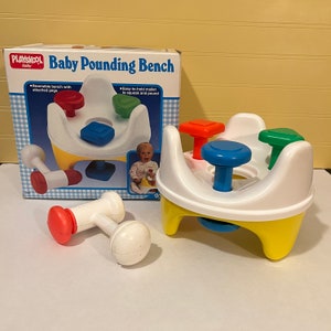 May include: A Playskool Baby Pounding Bench toy with a yellow base and white top. The bench has four colourful pegs that can be pounded with the included mallet. The box shows a baby sitting on the bench and playing with the toy.