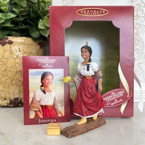 May include: A Hallmark Keepsake ornament from The American Girls Collection, featuring a figurine of Josefina. The figurine wears a red skirt, white blouse, and shawl. A card with Josefina's image and a small gold box are also present.