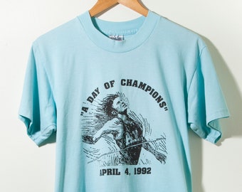 Puede incluir: Una camiseta azul claro con un gráfico en blanco y negro de una persona corriendo. El gráfico incluye el texto "A Day of Champions" y "April 4, 1992".
