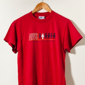 May include: Red short-sleeved t-shirt with a graphic print of the text "AWS" and a flame with five stick figures. The text below the graphic reads "We bring high tech to the human experience."