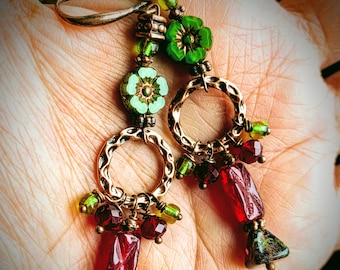 May include: Handmade earrings with copper-colored metal hoops and ear wires. The earrings feature green flower beads, red rectangular beads, and green and red accent beads. The design is intricate and detailed.