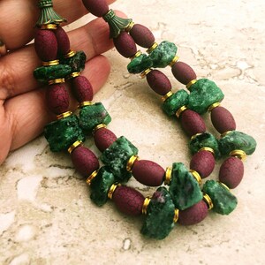 Può includere: Una collana a doppio filo con perline verdi e bordeaux. Le perline verdi sono di forma irregolare e sembrano essere fatte di pietra naturale. Le perline bordeaux sono di forma ovale e hanno una superficie testurizzata. La collana ha dettagli dorati.