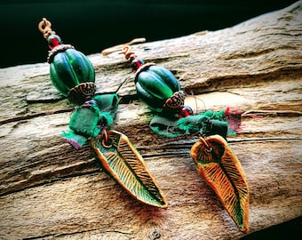 May include: A pair of dangle earrings with green glass beads, copper wire, and gold leaf accents. The earrings are hanging on a piece of driftwood.
