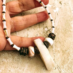 Peut inclure: Un collier de perles blanches et brunes avec un grand pendentif blanc. Le collier est composé de petites perles rondes et d'un pendentif rectangulaire plus grand. Le pendentif est fait d'un matériau blanc et a une surface lisse et polie.