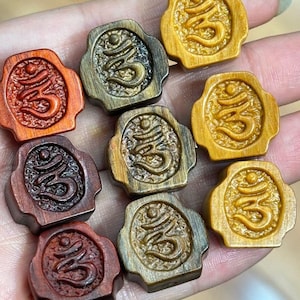 May include: A collection of nine wooden Om symbol pendants in various shades of brown and gold. Each pendant features a carved Om symbol within a decorative frame. The pendants are displayed in a hand.
