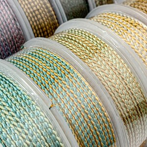 Può includere: Primo piano di diversi rocchetti di cordoncino intrecciato colorato in varie tonalità di verde, oro e beige. Le bobine sono disposte ordinatamente in fila, mostrando le diverse variazioni di colore e le trame del cordoncino. Il cordoncino sembra essere fatto di un materiale liscio e tessuto.