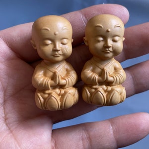 May include: Two small, wooden monk figurines. Each figurine is carved with closed eyes and hands clasped in prayer. The figures are light brown in color and have a smooth finish. The monks are seated with their legs crossed.