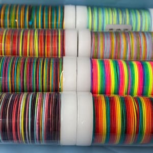 Puede incluir: Una colección de pulseras coloridas en varios tonos de rojo, naranja, amarillo, verde, azul, morado y rosa. Las pulseras están apiladas en tubos de plástico transparente con tapas blancas.
