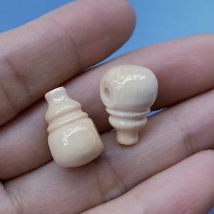 Könnte beinhalten: Zwei kleine, kunstvoll geschnitzte Knochenperlen in einer Hand. Eine Perle ist flaschenförmig, die andere rund mit einem kleinen Loch oben.