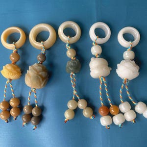 May include: Five decorative charms featuring smooth stone rings at the top, connected by multicolored braided cords to carved stone pendants in shapes of flowers and rounded figures, finished with small dangling stone beads at the ends.