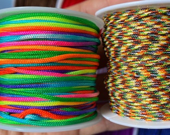 Puede incluir: Dos carretes de cuerda de colores. Un carrete tiene un arcoíris de colores, incluyendo verde, naranja, rosa y morado. El otro carrete tiene un patrón de camuflaje con verde, marrón y blanco.
