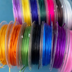 May include: Spools of colorful elastic cord in various shades, including yellow, orange, green, black, blue, purple, pink, and red. The cords are wound on clear plastic spools, ideal for crafting and jewelry making.