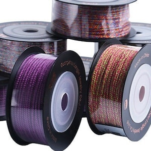 Puede incluir: Una colección de carretes de hilo de bordar de colores, cada uno enrollado alrededor de un carrete de plástico negro con una ventana de plástico transparente. Los carretes están apilados uno encima del otro, con el carrete superior siendo un hilo multicolor.