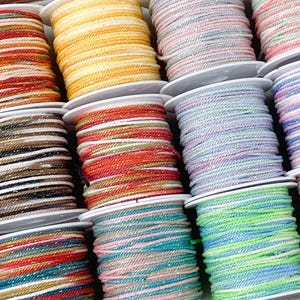 Pode incluir: Uma bandeja de madeira contém rolos de cordão colorido em várias tonalidades, incluindo laranja, amarelo, rosa, verde e azul. Cada rolo é cuidadosamente enrolado, oferecendo uma variedade de opções para artesanato e projetos DIY.
