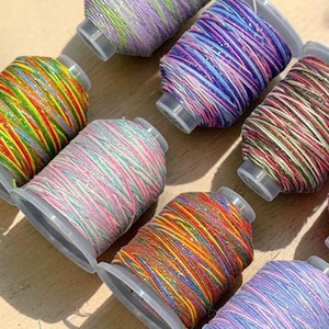 May include: A collection of eleven spools of multi-colored thread arranged in a diagonal pattern. Each spool features a unique combination of vibrant hues, including shades of blue, pink, orange, green, and purple, with a subtle metallic shimmer.