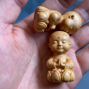 May include: Three small, wooden monk figurines. One is seated with hands in prayer, eyes closed. Two others are in a fetal position. The figurines are light brown and appear to be carved from a single piece of wood.