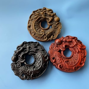 K&ouml;nnte beinhalten: Drei Holz-Drachen-Anh&auml;nger, jeder in einer anderen Farbe geschnitzt: braun, schwarz und rot. Die Drachen sind um eine kreisf&ouml;rmige &Ouml;ffnung gekr&uuml;mmt, mit dem Kopf nach au&szlig;en.