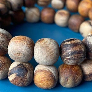 May include: A wooden beaded bracelet with round beads in natural tones of brown, tan, and cream. The bracelet is displayed on a blue surface. The beads have a diameter of approximately 1 cm. The bracelet's natural colours are highlighted by the blue background.