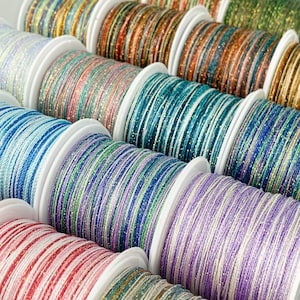 May include: Close-up of several spools of colorful, glittery thread. The thread is wound on white spools and comes in various colors, including blue, green, pink, and gold. The thread appears to be made of a shiny, metallic material.