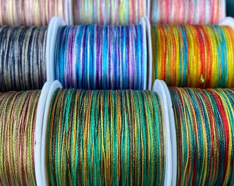May include: Close-up of spools of colourful embroidery floss. The floss is wound around white plastic spools and features a variety of colours and patterns, including green, blue, red, yellow, and brown.