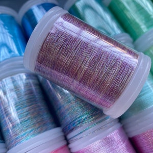 Può includere: Primo piano di diversi rocchetti di filo metallico colorato. Il filo è disponibile in varie tonalità di blu, rosa e verde, con una qualità brillante e iridescente. Ogni rocchetto è racchiuso in un contenitore di plastica trasparente con un coperchio bianco. Per ricamo.