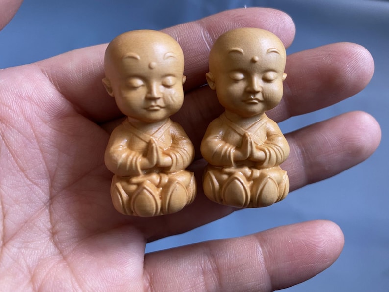 May include: Two small, wooden monk figurines. Each figurine is carved with closed eyes and hands clasped in prayer. The figures are light brown in color and have a smooth finish. The monks are seated with their legs crossed.