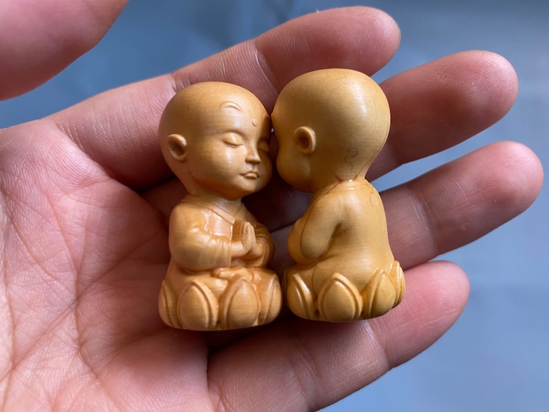 May include: Two small, wooden figurines of meditating monks. The monks are light brown, with closed eyes and hands clasped in prayer. They are seated on lotus flower bases, with one monk facing the other.