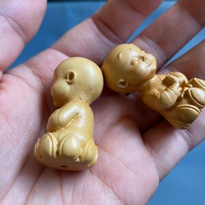 May include: Two small, light-colored wooden figurines of seated babies. One is upright, the other is lying down. The babies have rounded heads and are in a meditative pose. The figurines are carved with details.