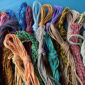 Può includere: Una collezione di cordini intrecciati in vari colori, tra cui arancione, blu, rosso, giallo, verde e oro metallizzato. Ogni cordino è avvolto e annodato, con fili multicolori intrecciati insieme per creare un aspetto strutturato simile a una corda.